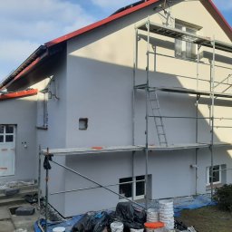 Bourne - Dom po tynkowaniu elewacji, widoczne rusztowania i przygotowane farby. Świeży tynk w odcieniach szarości, czerwony dach. Prace w toku, widoczny pracownik w tle.