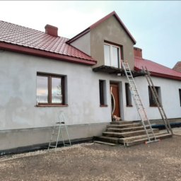 Bourne - Dom jednorodzinny w trakcie prac elewacyjnych: ściany przygotowane pod tynk, widoczne drabiny, czerwony dach. Ujęcie z dołu, w tle pochmurne niebo.