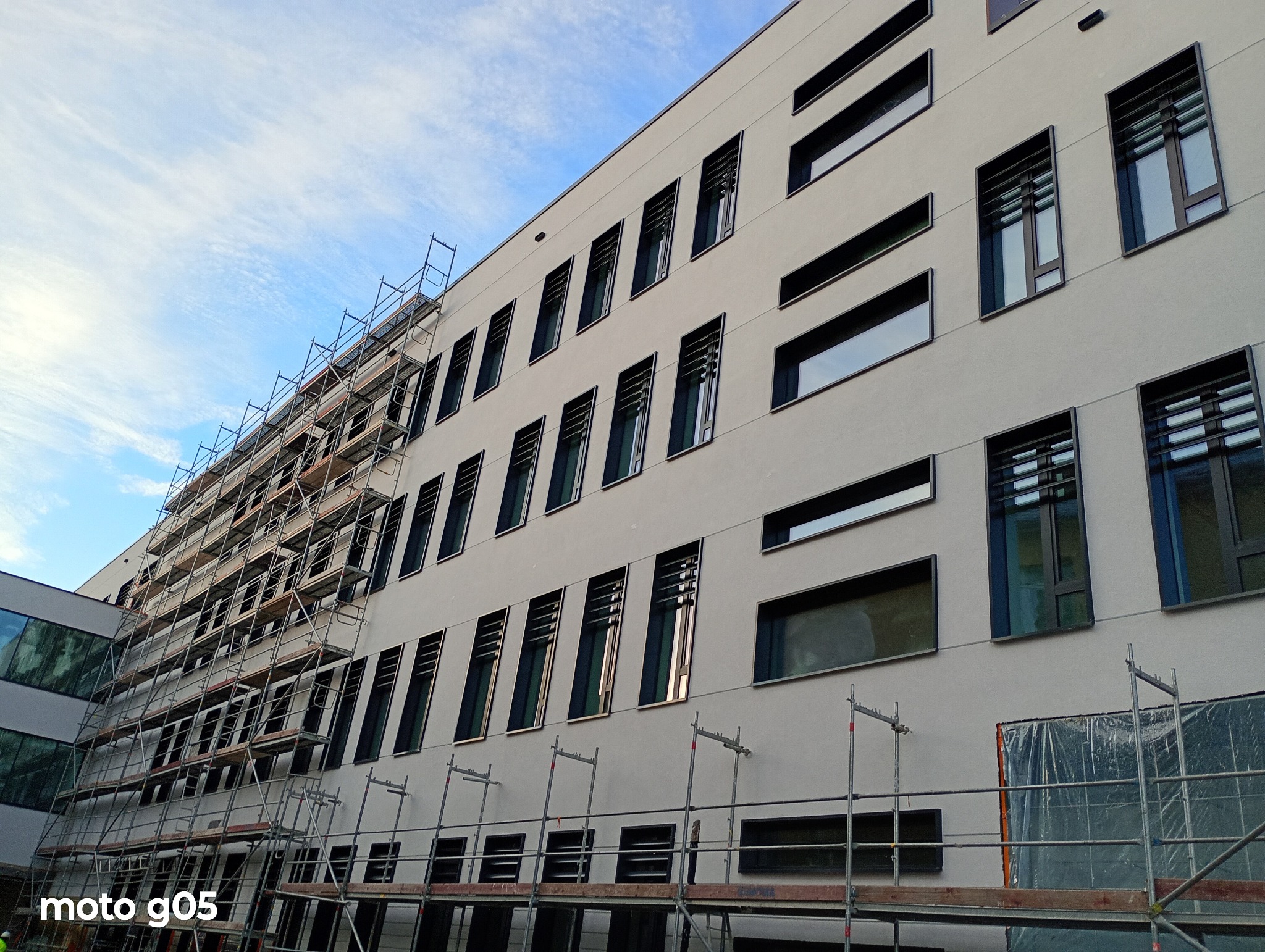 Elewacja budynku w trakcie prac wykończeniowych, widoczne rusztowania i świeżo otynkowana ściana z oknami. Błękitne niebo w tle.