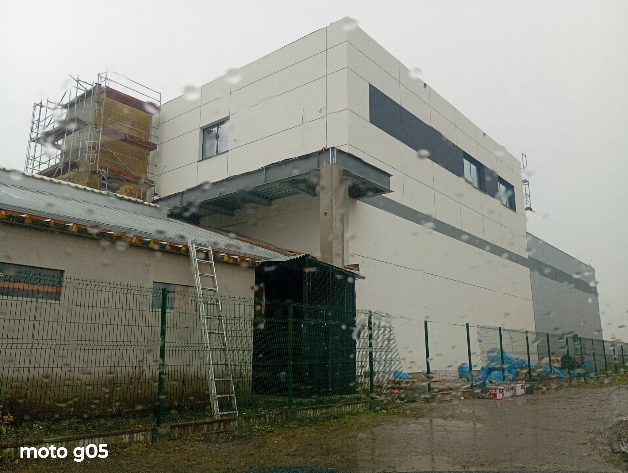 Ocieplanie elewacji budynku w trakcie realizacji. Widoczne rusztowania, materiały izolacyjne i fragmenty elewacji z panelami. Ujęcie zza szyby z kroplami deszczu.