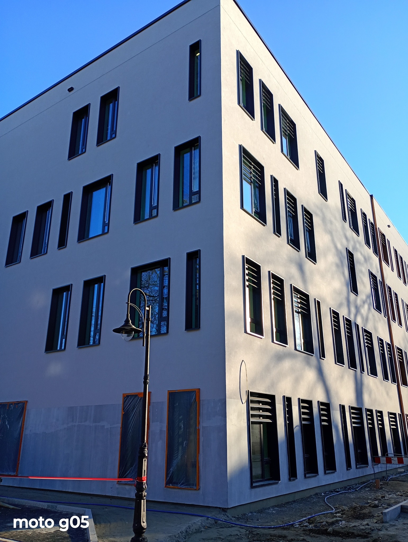 Nowoczesny budynek z szarą elewacją i ciemnymi ramami okiennymi. Część okien zabezpieczona folią. Widoczna latarnia uliczna i fragment placu budowy.