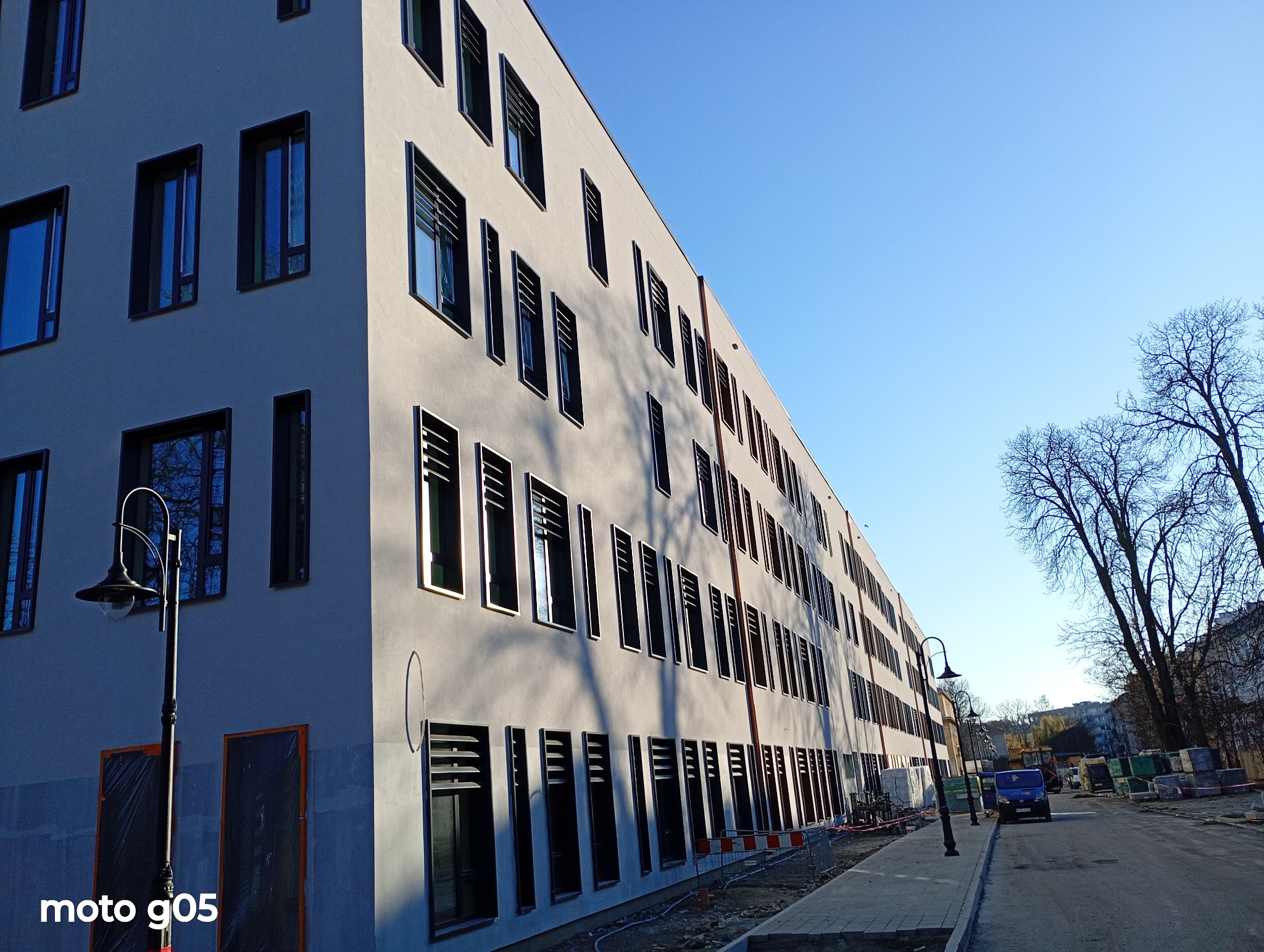 Nowoczesny budynek z szarą elewacją i regularnym układem okien z żaluzjami, widziany z perspektywy ulicy w słoneczny dzień. Widoczne lampy uliczne i drzewa.