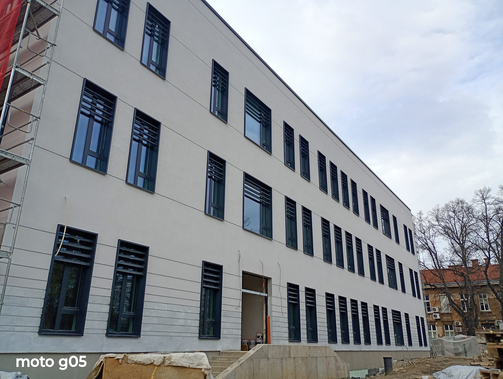 Trzypiętrowy budynek z szarą elewacją i ciemnymi oknami z żaluzjami, widziany pod kątem. Fragment rusztowania po lewej, teren budowy u dołu. Pochmurne niebo w tle.