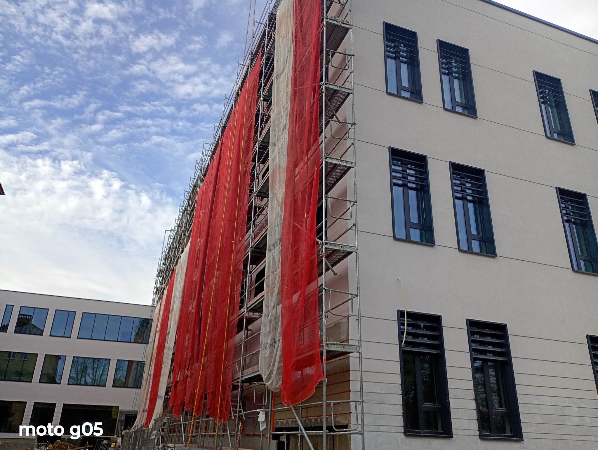 Elewacja budynku w trakcie prac w Krakowie, osłonięta siatką na rusztowaniach. Widoczne okna z ciemnymi ramami i fragment drugiego budynku w tle.