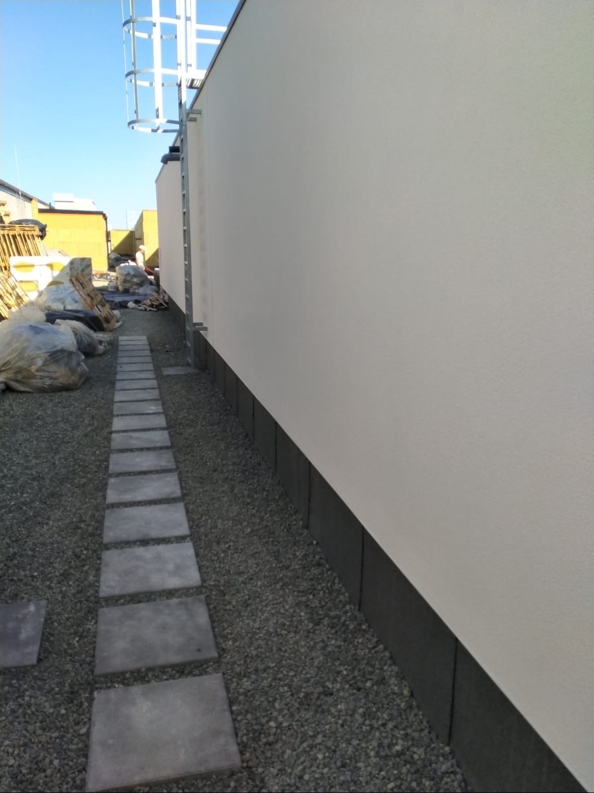 Gładka, jasna elewacja budynku z ciemną podmurówką, widoczna z perspektywy ścieżki z płyt. Metalowa drabina na ścianie, gruz i materiały budowlane w tle.