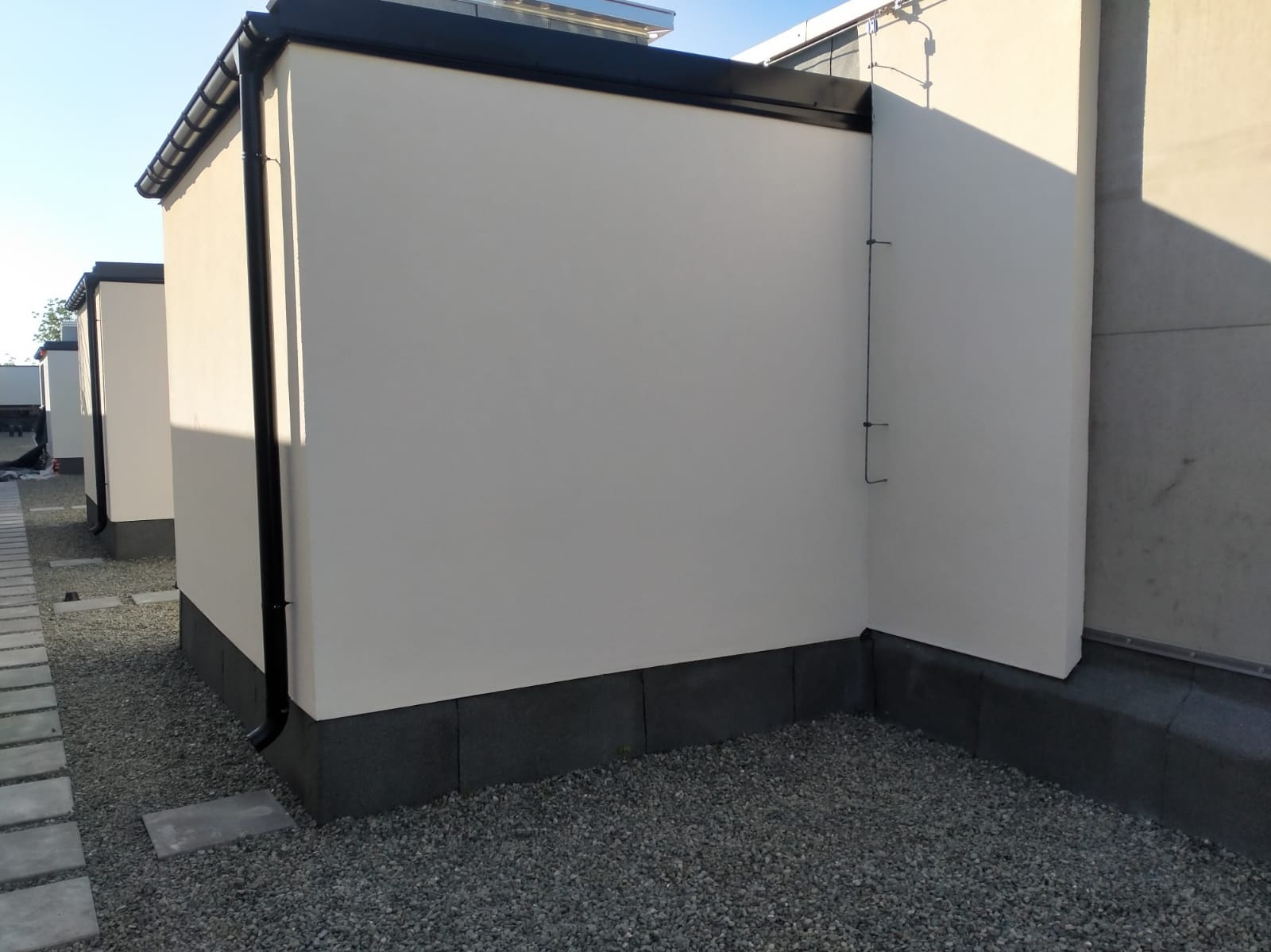 Biała elewacja budynku z czarnym orynnowaniem, widok z poziomu gruntu na szary żwir i fragment chodnika z płyt. Minimalistyczny styl, jasne oświetlenie.