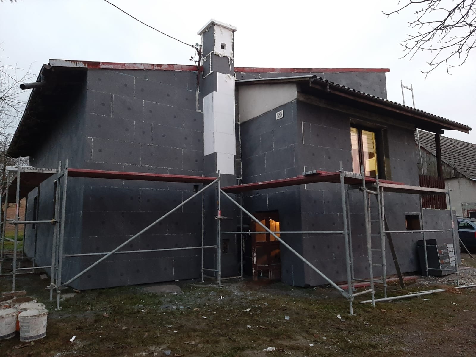 Dom w trakcie ocieplania, pokryty szarymi płytami izolacyjnymi. Widoczne rusztowania i fragment ocieplonego komina. Prace w toku, surowy krajobraz.