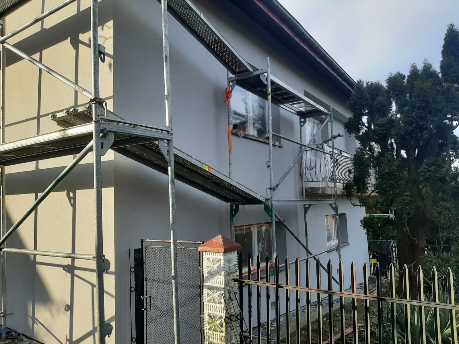 Świeżo otynkowana elewacja domu w kolorze szarości, z widocznymi rusztowaniami i zabezpieczonymi oknami folią. Fragment metalowego ogrodzenia na pierwszym planie.