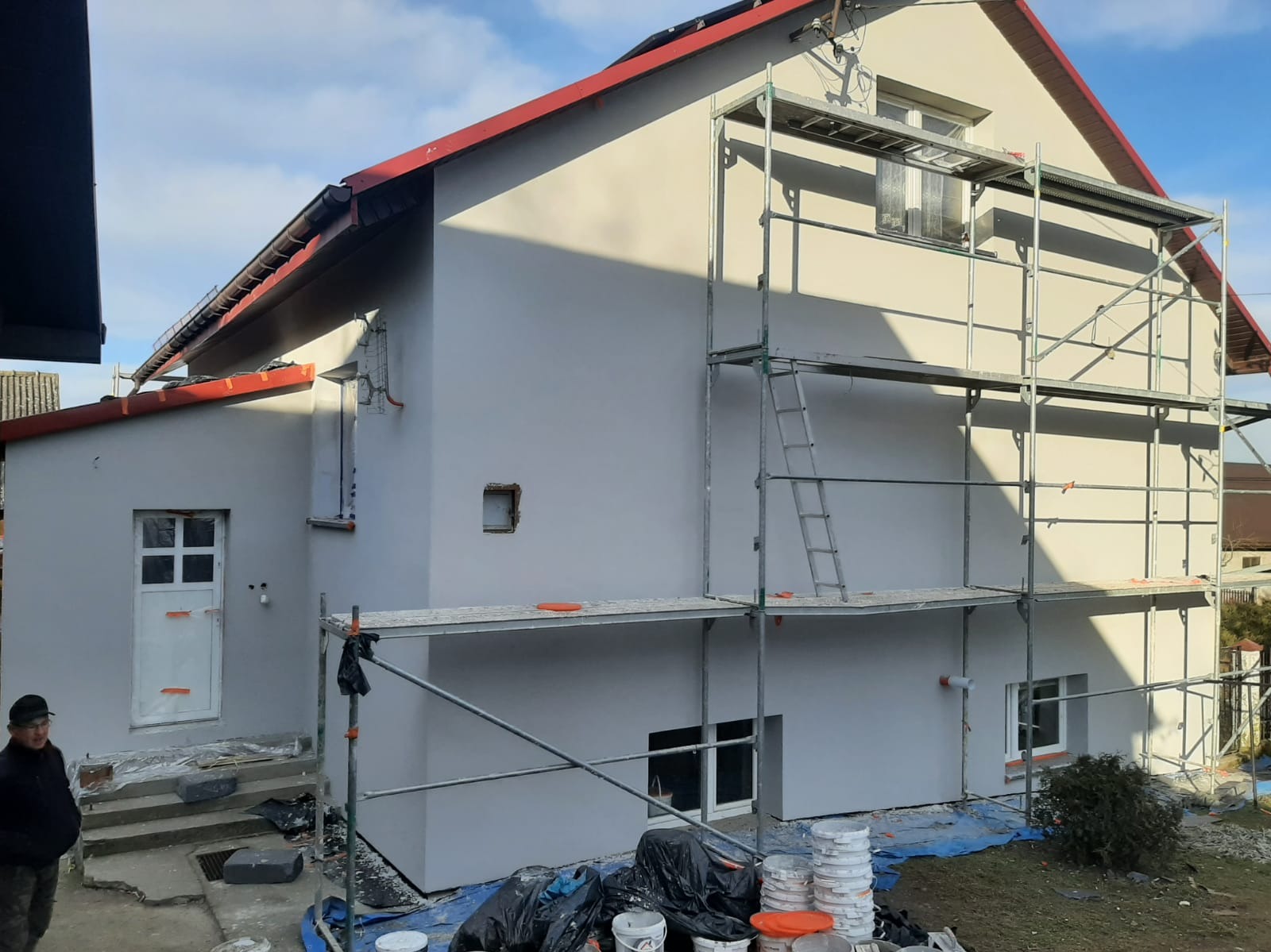 Dom po tynkowaniu elewacji, widoczne rusztowania i przygotowane farby. Świeży tynk w odcieniach szarości, czerwony dach. Prace w toku, widoczny pracownik w tle.