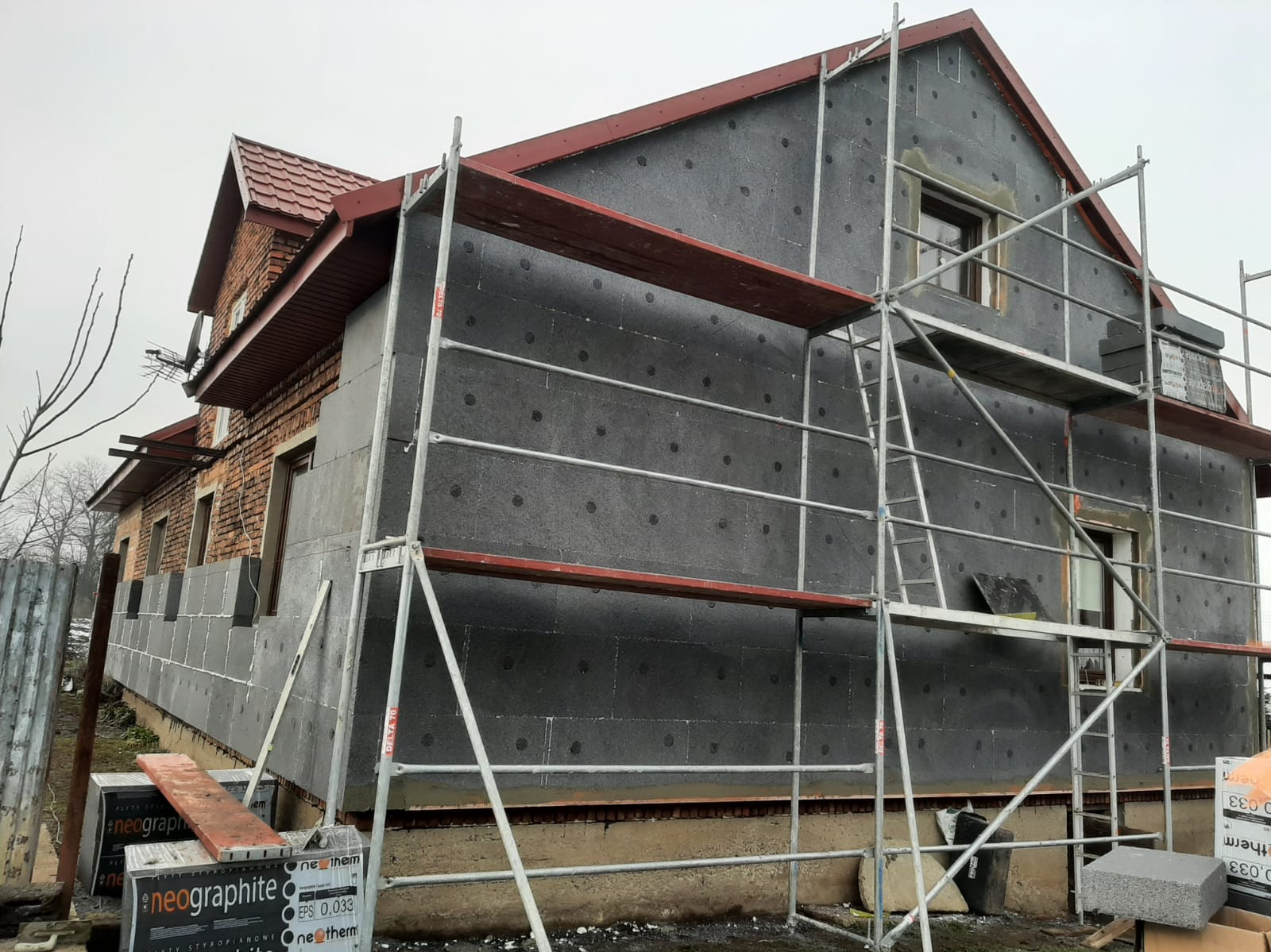 Dom w trakcie ocieplania grafitowym styropianem, widoczne rusztowania i częściowo ocieplona elewacja. Prace na wysokości.