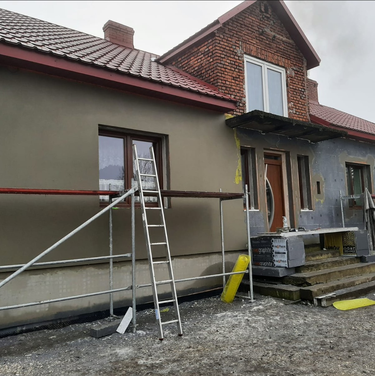 Dom w trakcie ocieplania elewacji: część ściany otynkowana na szaro, rusztowanie, styropian grafitowy, fragment ściany z ociepleniem. Widoczne okna i drzwi.
