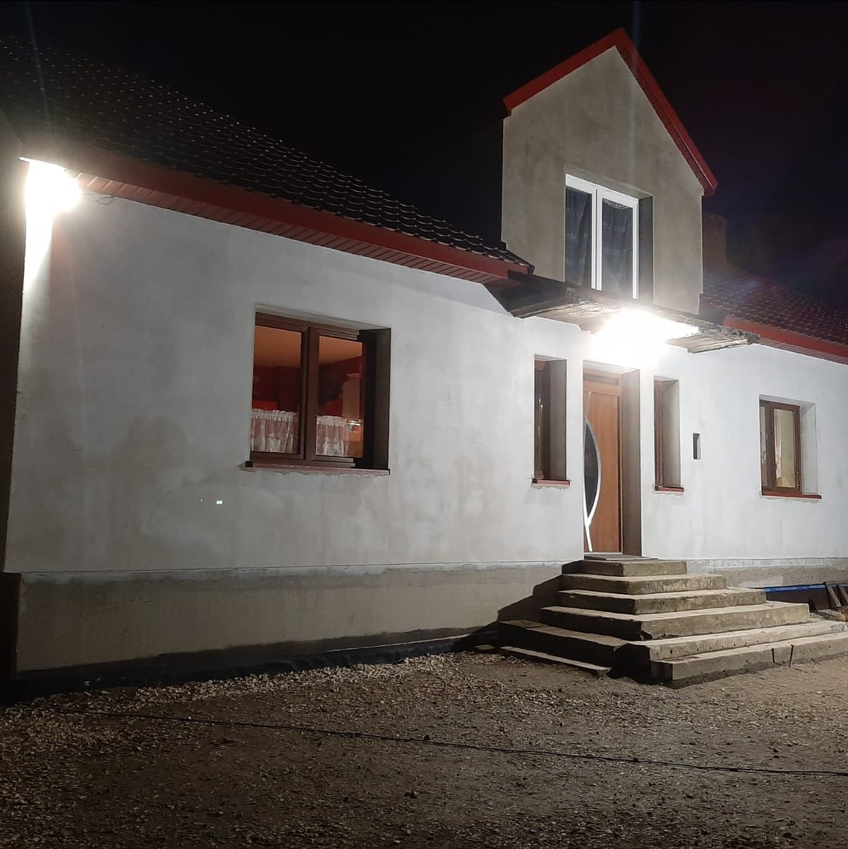Nocne ujęcie domu w trakcie prac elewacyjnych: surowy tynk, widoczne schody wejściowe, okna z drewnianą ramą i czerwone wykończenia dachu. Oświetlenie podkreśla fakturę.