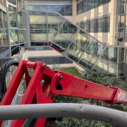 Langteam - Czerwony podnośnik koszowy na tle nowoczesnego biurowca z przeszkloną fasadą i zielonym patio. Widok z góry, błękitne niebo z chmurami odbija się w oknach.