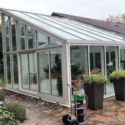 Langteam - Szklana oranżeria przylegająca do domu, po umyciu. Widoczny wózek ze sprzętem do mycia okien, donice z kwiatami i mokra kostka brukowa. Jesienne liście na ziemi.