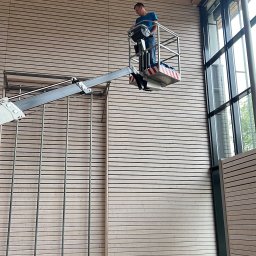 Langteam - Pracownik na podnośniku koszowym czyści wysoką, drewnianą ścianę w hali sportowej. Widoczne duże okna i drewniana konstrukcja dachu.