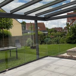 Langteam - Szklana zabudowa tarasu z widokiem na ogród. Jasne płytki na podłodze, nowoczesna konstrukcja z szarymi profilami. Widoczne ślady po umyciu na płytkach.