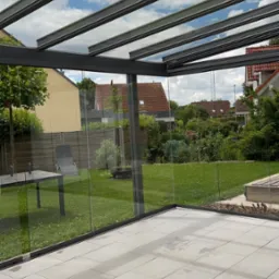 Szklana zabudowa tarasu z widokiem na ogród. Jasne płytki na podłodze, nowoczesna konstrukcja z szarymi profilami. Widoczne ślady po umyciu na płytkach.