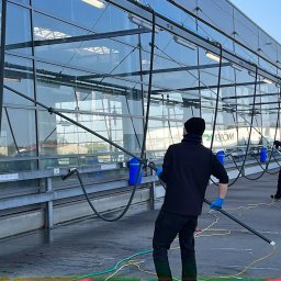 Langteam - Dwóch mężczyzn w trakcie mycia szklanej fasady budynku za pomocą specjalistycznych lanc i węży. Widoczne niebieskie pojemniki i zaparkowany samochód.