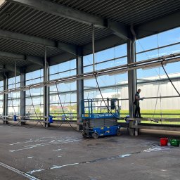 Langteam - Mycie okien hali przemysłowej z użyciem podnośnika i drabiny. Widoczne ślady wody na posadzce, błękitne niebo za oknami i pracownik w trakcie czyszczenia.