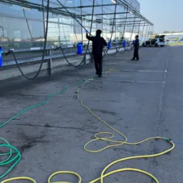 Dwaj pracownicy myją szklaną konstrukcję z użyciem długich lanc i węży. Widoczne niebieskie wiadra i zaparkowany samochód w tle. Praca na zewnątrz w słoneczny dzień.