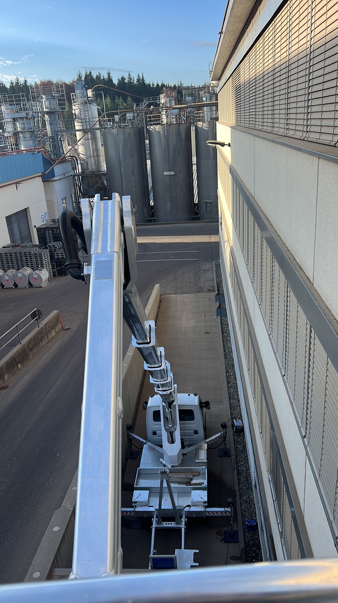 Podnośnik koszowy na tle przemysłowego budynku z metalowymi zbiornikami. Widok z góry, z perspektywy kosza. Jasne światło słoneczne.