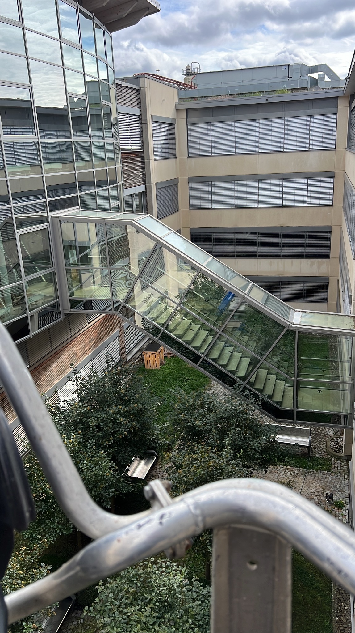 Szklany łącznik między budynkami biurowymi, widok z góry z elementami rusztowania w kadrze. Zieleń wewnątrz łącznika i na dziedzińcu. Architektura biurowa.