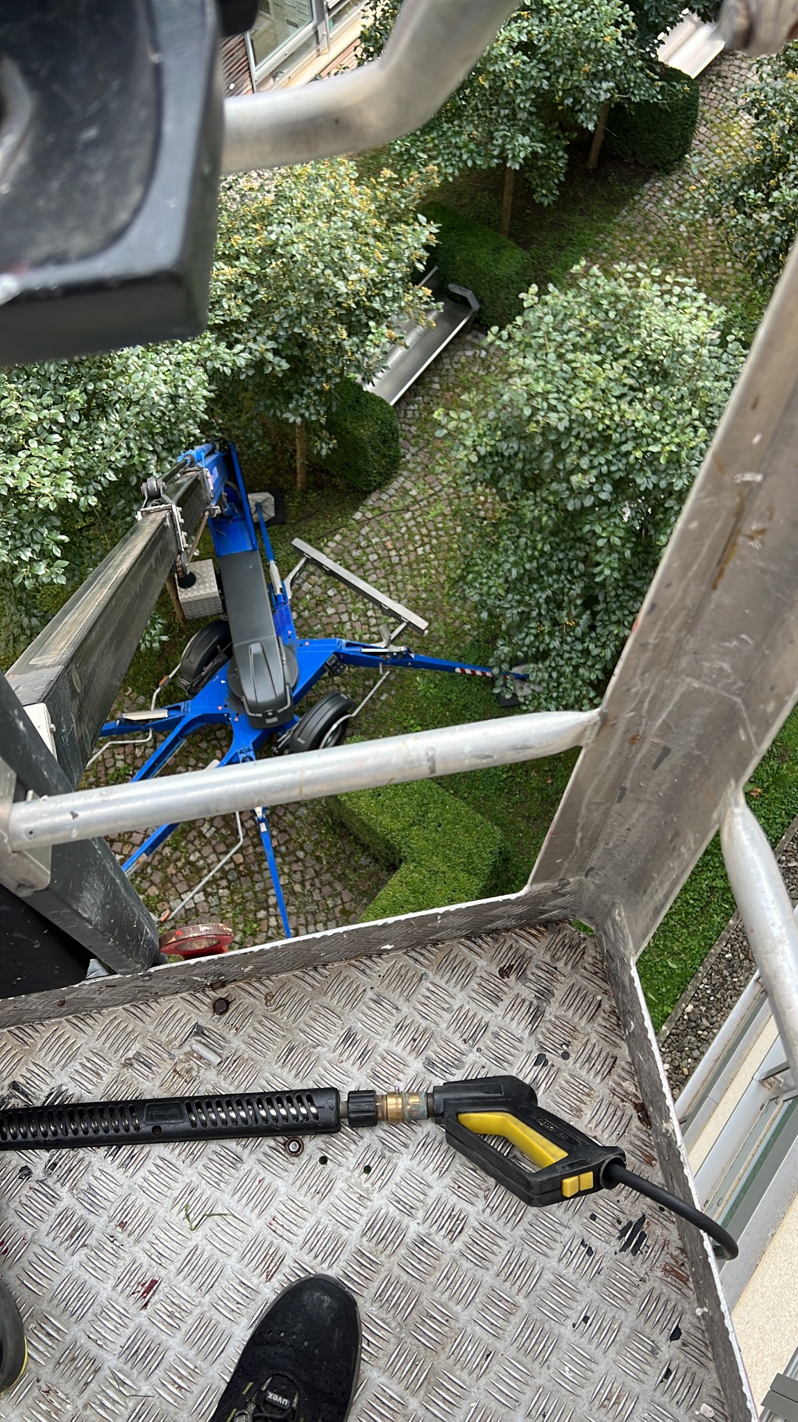 Widok z podnośnika koszowego na kostkę brukową i narzędzie do mycia ciśnieniowego. W tle zieleń i fragment budynku. Perspektywa z góry.