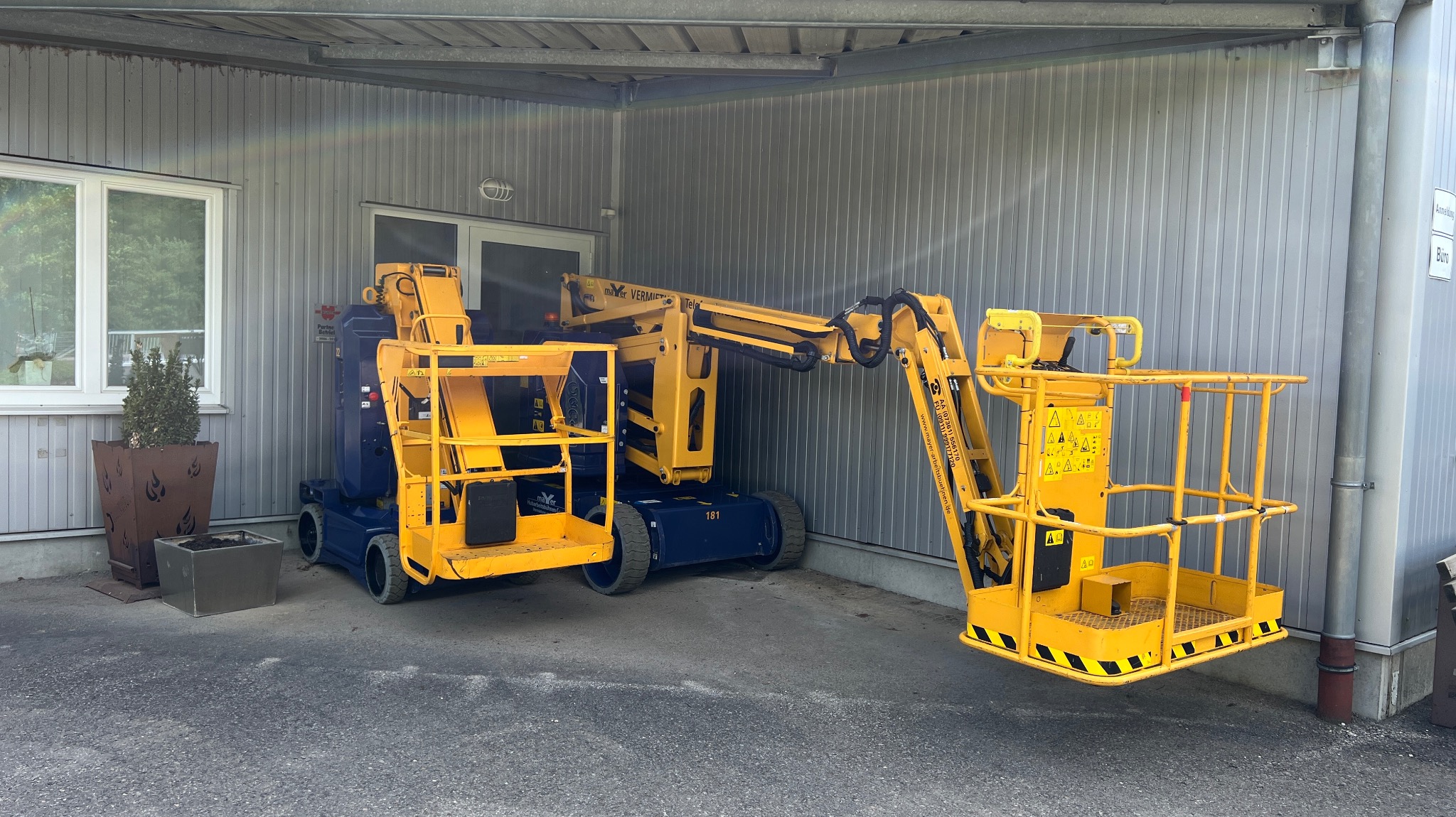 Żółto-niebieski podnośnik koszowy marki Mayer Vermietet, zaparkowany przed budynkiem z szarej blachy falistej. Widoczne dwie platformy robocze i elementy konstrukcyjne.