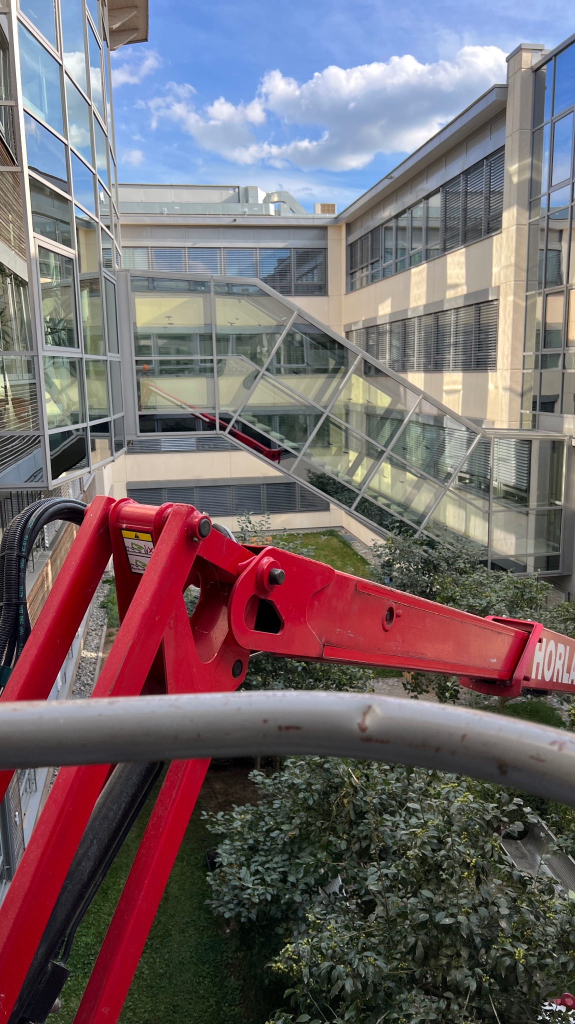 Czerwony podnośnik koszowy na tle nowoczesnego biurowca z przeszkloną fasadą i zielonym patio. Widok z góry, błękitne niebo z chmurami odbija się w oknach.