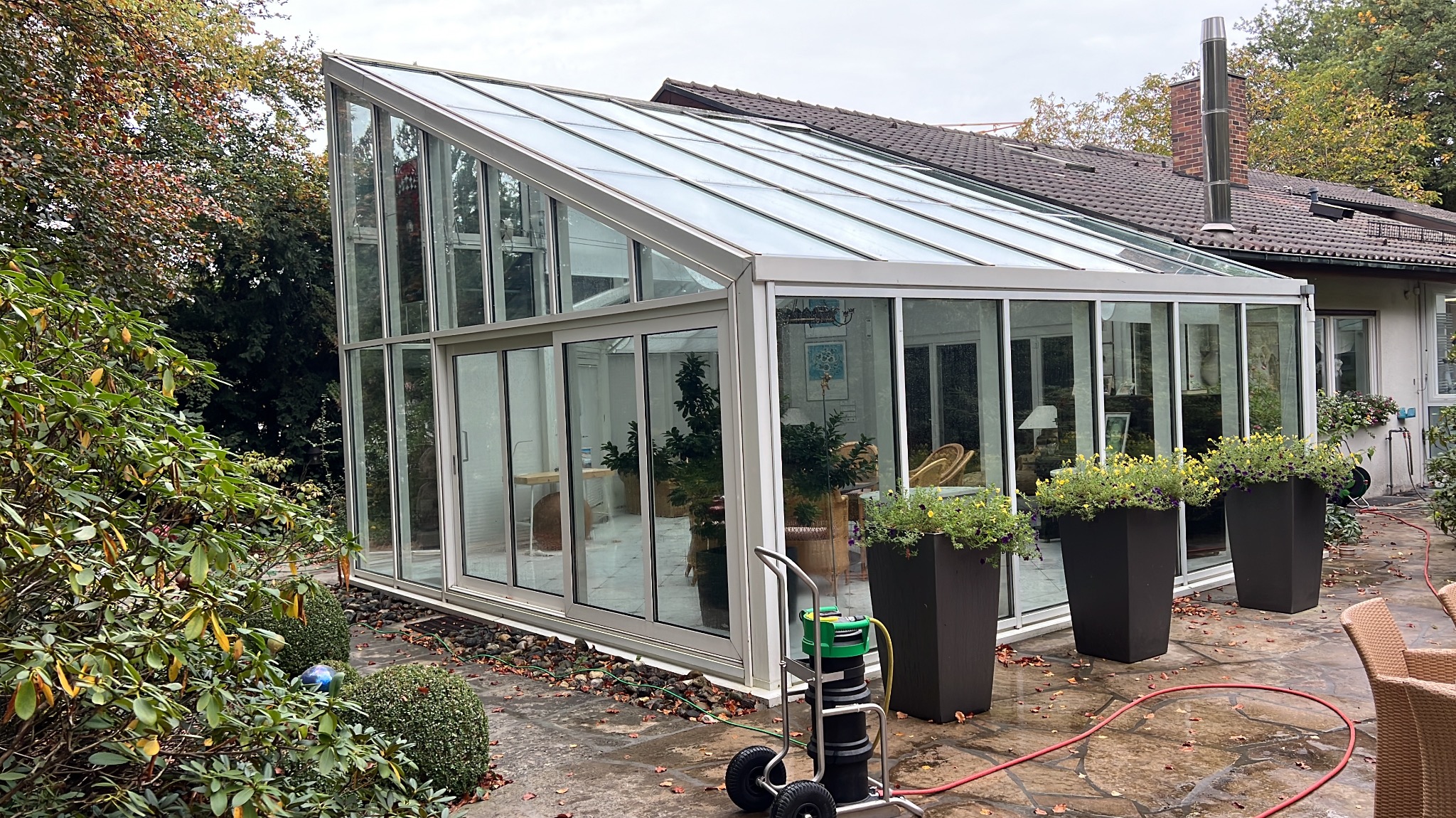 Szklana oranżeria przylegająca do domu, po umyciu. Widoczny wózek ze sprzętem do mycia okien, donice z kwiatami i mokra kostka brukowa. Jesienne liście na ziemi.