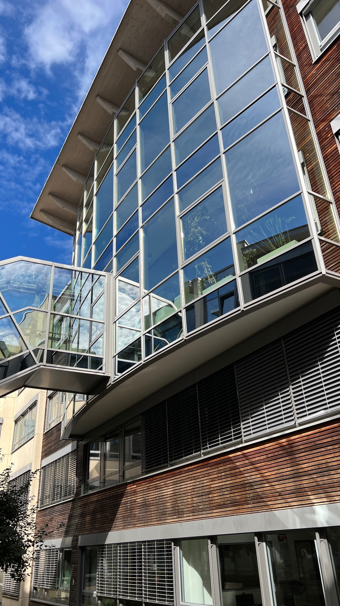 Nowoczesny budynek biurowy z przeszkloną fasadą i łącznikiem, odbijający błękitne niebo. Widoczne drewniane elementy elewacji i żaluzje okienne.