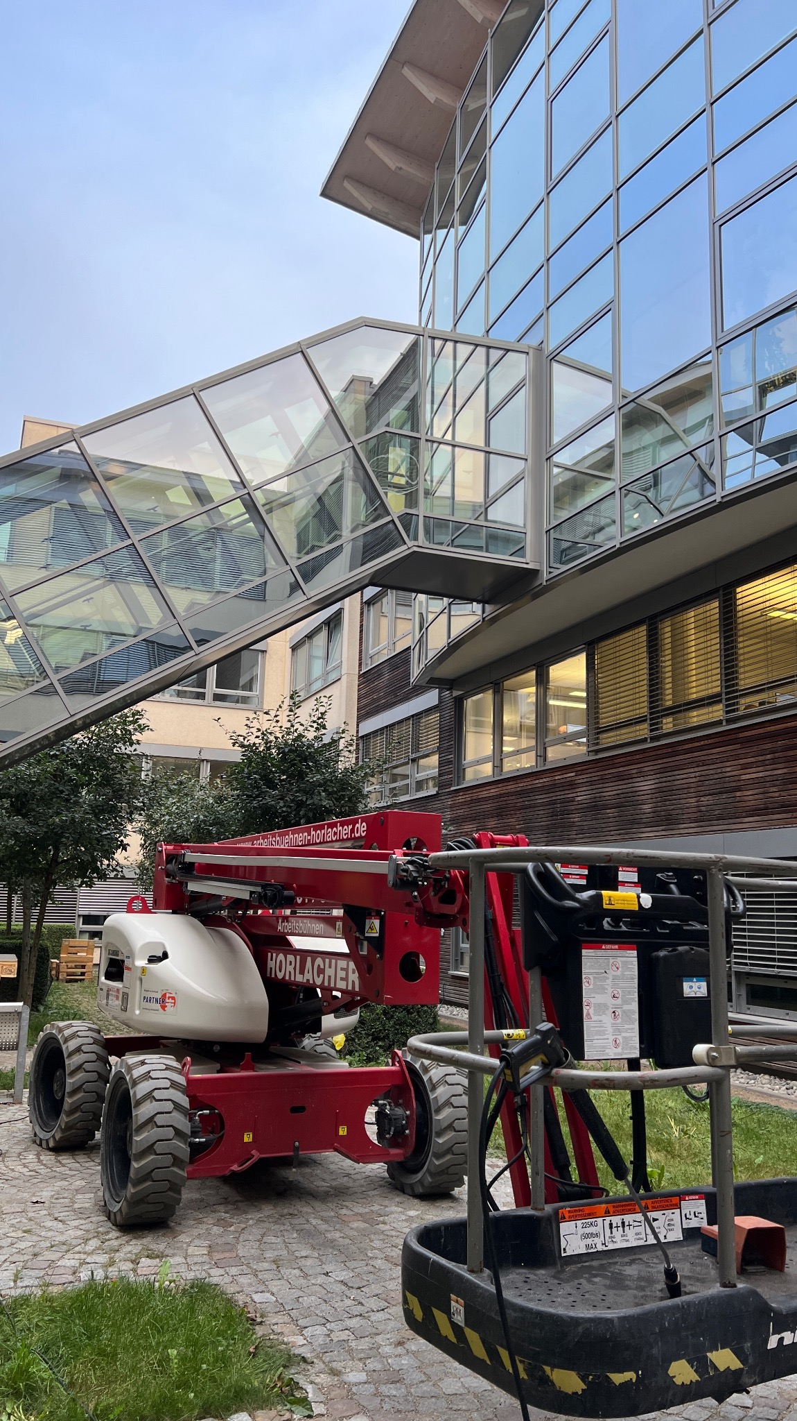 Czerwony podnośnik koszowy Horlacher przed nowoczesnym budynkiem z przeszklonym łącznikiem. Widoczne okna i kostka brukowa.