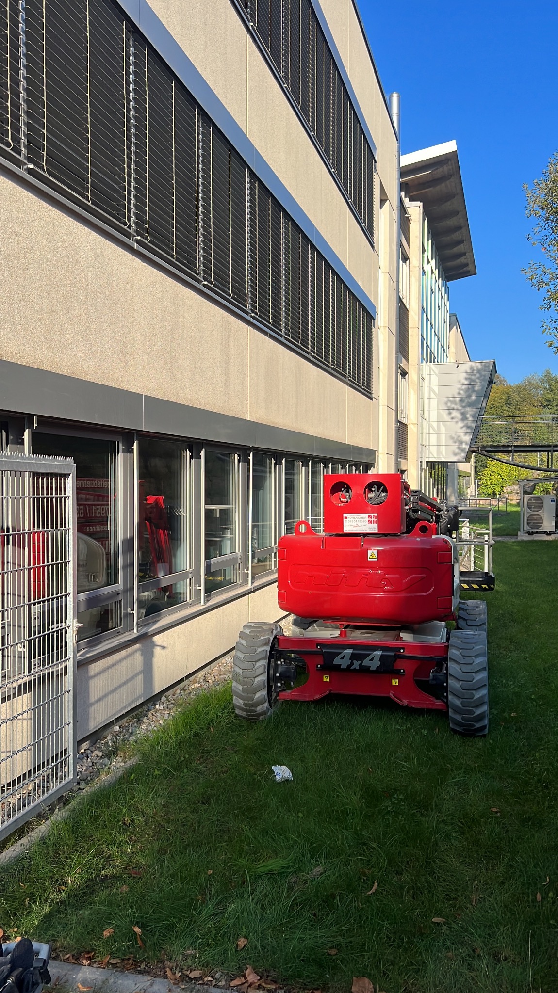 Czerwony podnośnik koszowy 4x4 stoi na trawie przed budynkiem z oknami i żaluzjami. Widoczne elementy konstrukcyjne budynku i zieleń w tle.