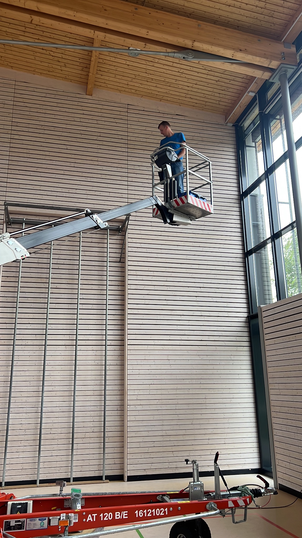 Pracownik na podnośniku koszowym czyści wysoką, drewnianą ścianę w hali sportowej. Widoczne duże okna i drewniana konstrukcja dachu.