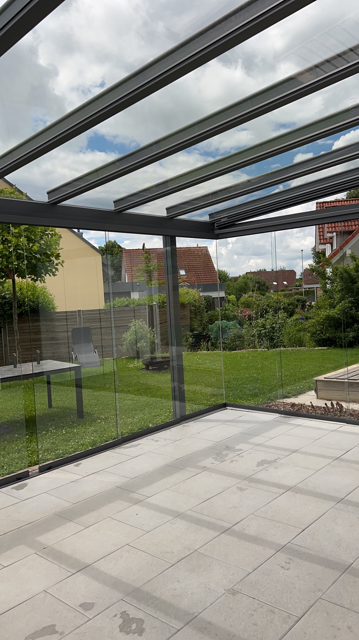Szklana zabudowa tarasu z widokiem na ogród. Jasne płytki na podłodze, nowoczesna konstrukcja z szarymi profilami. Widoczne ślady po umyciu na płytkach.