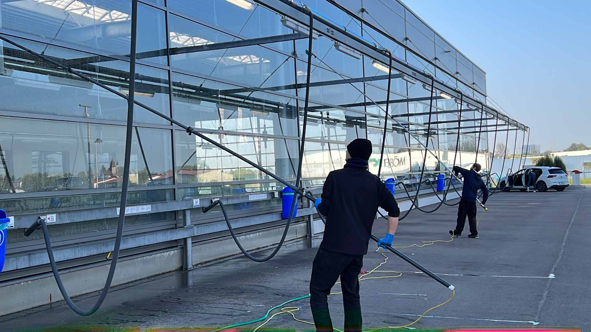 Dwóch mężczyzn w trakcie mycia szklanej fasady budynku za pomocą specjalistycznych lanc i węży. Widoczne niebieskie pojemniki i zaparkowany samochód.