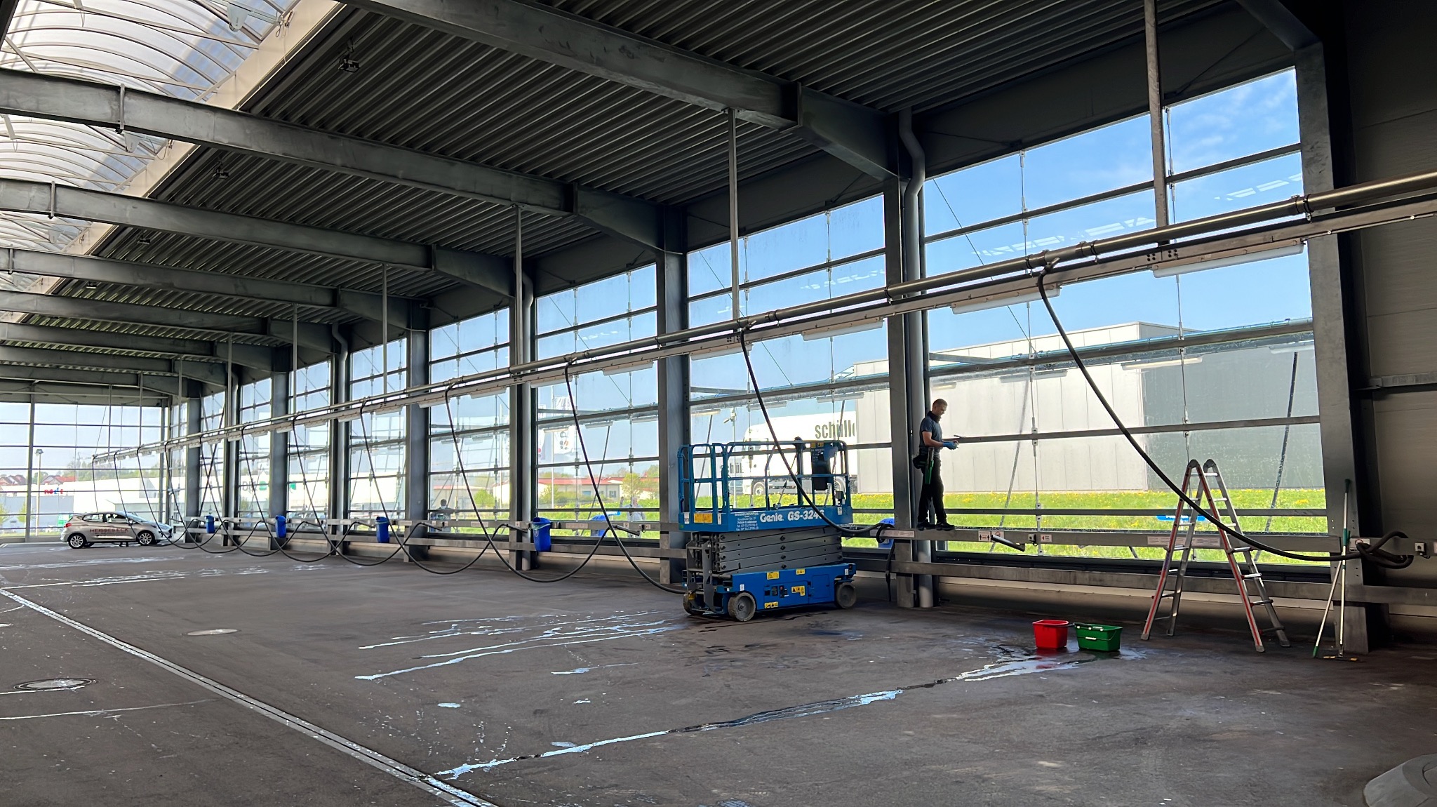 Mycie okien hali przemysłowej z użyciem podnośnika i drabiny. Widoczne ślady wody na posadzce, błękitne niebo za oknami i pracownik w trakcie czyszczenia.