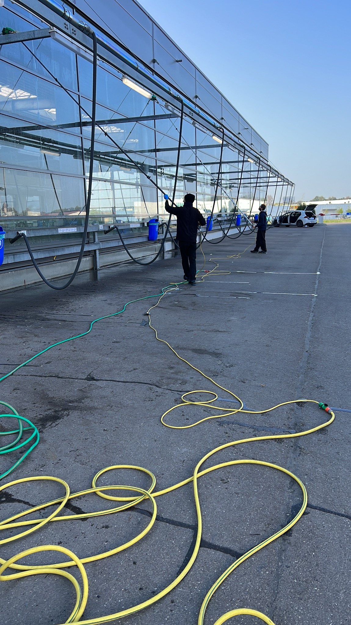 Dwaj pracownicy myją szklaną konstrukcję z użyciem długich lanc i węży. Widoczne niebieskie wiadra i zaparkowany samochód w tle. Praca na zewnątrz w słoneczny dzień.