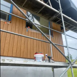 Elewacja domu w trakcie malowania z rusztowania. Widoczne narzędzia, puszka farby i zabezpieczone okna. Drewniana okładzina elewacyjna w odcieniach brązu.