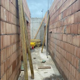 Dorinex-Budownictwo - Wąski kadr na wznoszone ściany z czerwonej cegły ceramicznej, z widocznymi deskami szalunkowymi i fragmentem betonowej konstrukcji w tle, w pochmurny dzień.