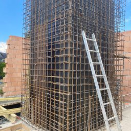 Dorinex-Budownictwo - Zbrojenie pionowego elementu konstrukcyjnego z deskowaniem i drabiną na budowie. Widoczne cegły i błękitne niebo w tle. Detal konstrukcji żelbetowej.