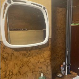 Dorinex-Budownictwo - Ukończona łazienka z brązowymi płytkami imitującymi marmur, białą umywalką i lustrem w białej ramie. Widoczne rury i akcesoria łazienkowe.