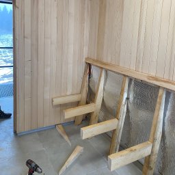 Dorinex-Budownictwo - Montaż boazerii drewnianej na ścianie z izolacją, widoczny szkielet konstrukcji i wkrętarka. Fragment widoku na zewnątrz przez okno. Prace wykończeniowe wewnątrz budynku.