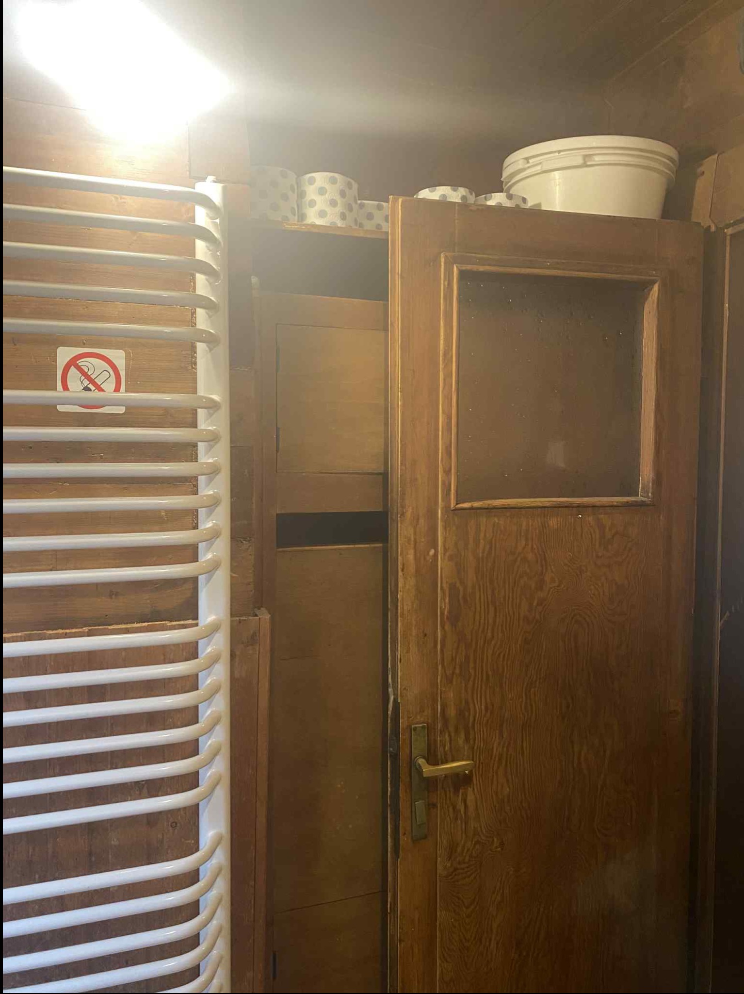 Biały grzejnik łazienkowy na tle drewnianej ściany, obok drewniane drzwi z szybą i zakaz palenia. Na szafce rolki papieru toaletowego i białe wiadro.