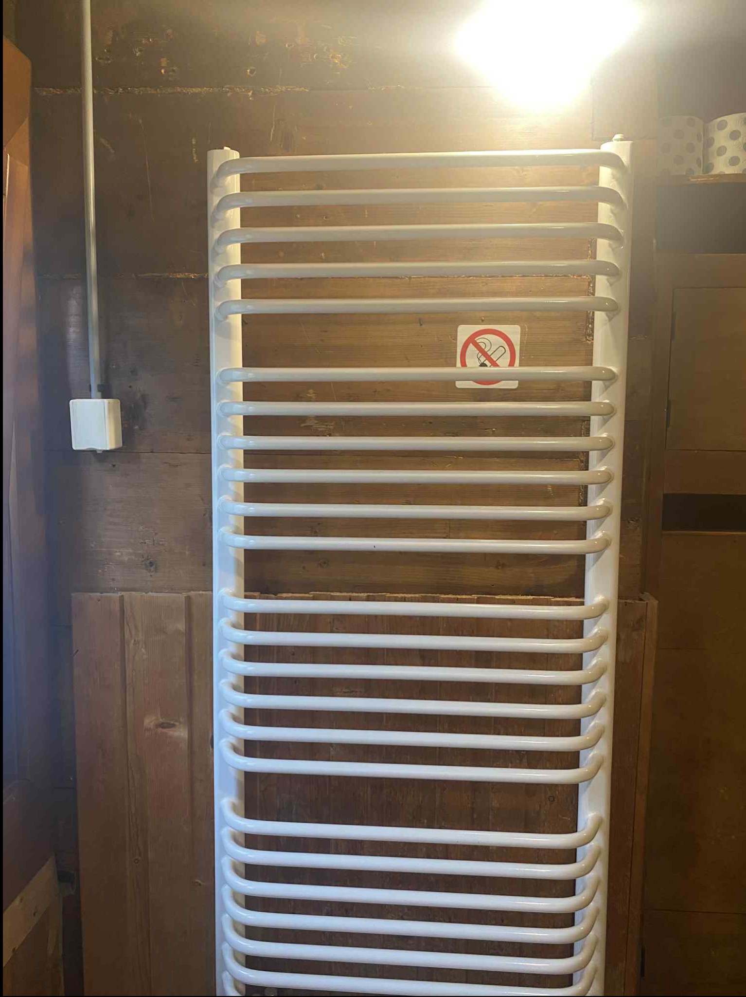 Biały, drabinkowy grzejnik łazienkowy na tle boazerii. Na grzejniku naklejka z przekreślonym papierosem. Widoczna instalacja elektryczna i fragment szafki.