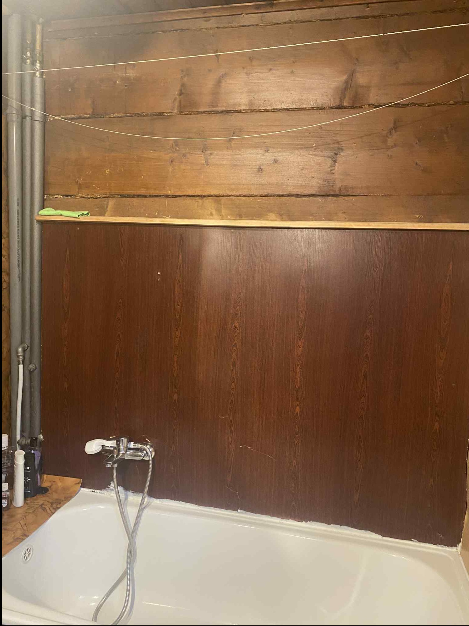 Wykończona ściana w łazience z brązową okładziną imitującą drewno nad wanną. Widoczne rury i armatura. Górna część ściany wykończona drewnianymi deskami.