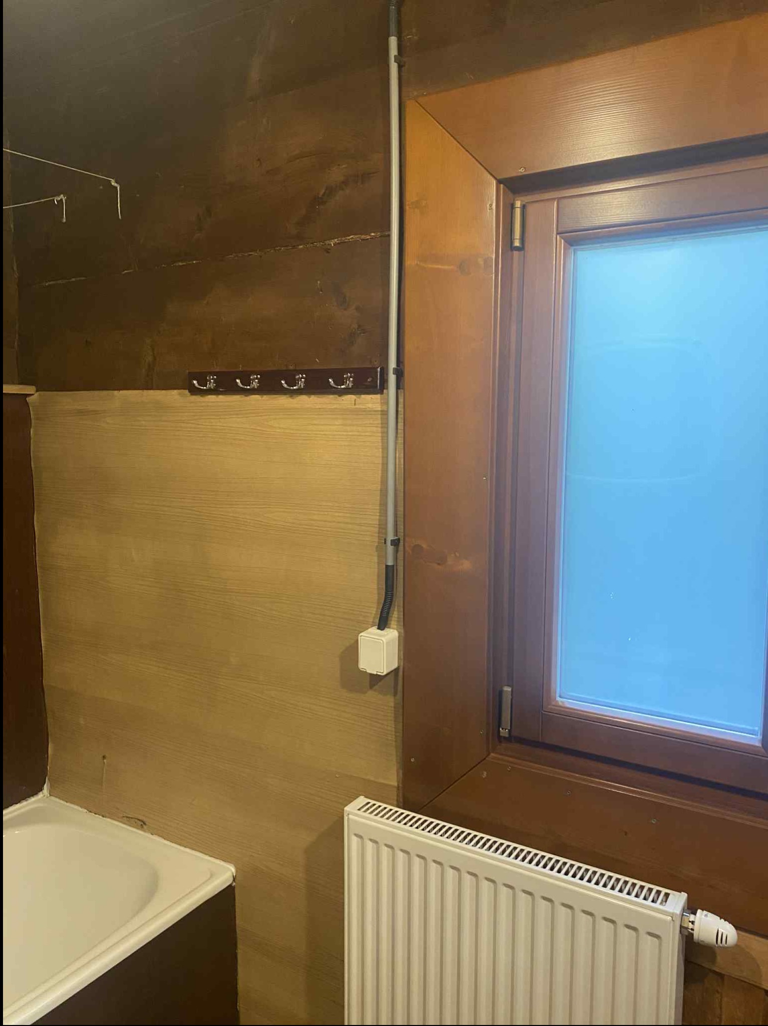 Drewniane panele ścienne w łazience z grzejnikiem i oknem w drewnianej ramie. Widoczna instalacja elektryczna. Haczyki na ścianie.
