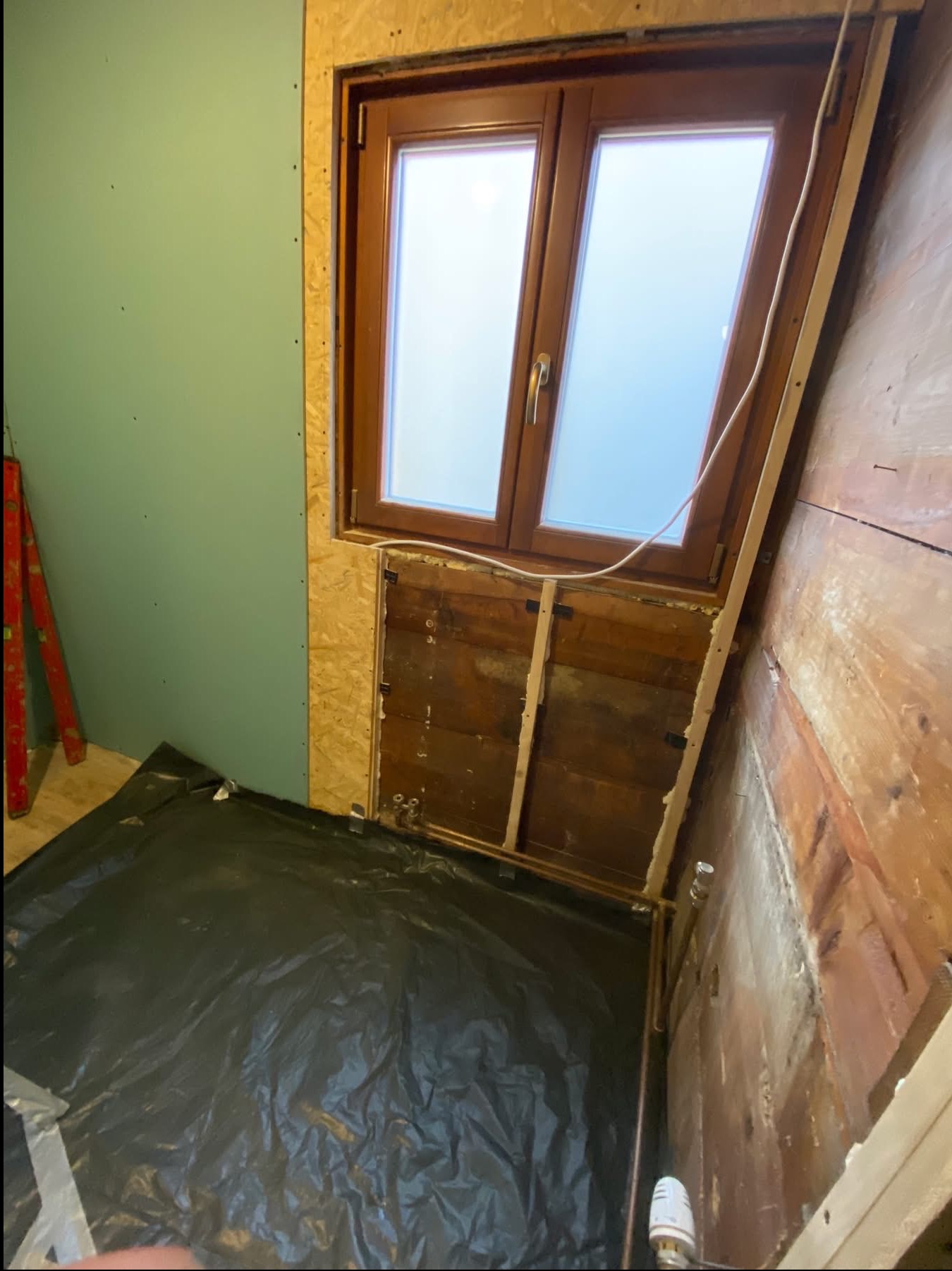 Wnętrze w trakcie remontu: okno, fragment ściany z płytą karton-gips, odsłonięta drewniana ściana, rury i folia ochronna na podłodze. Widoczne elementy instalacji.