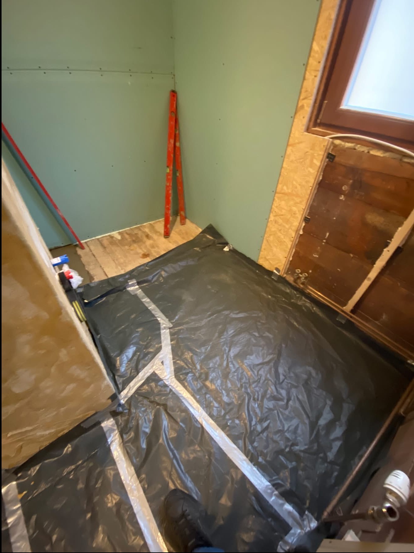 Przygotowanie podłoża: folia izolacyjna z taśmą na drewnianej podłodze, ściany z płyt karton-gips, poziomica oparta o ścianę. Widoczne rury instalacji grzewczej.