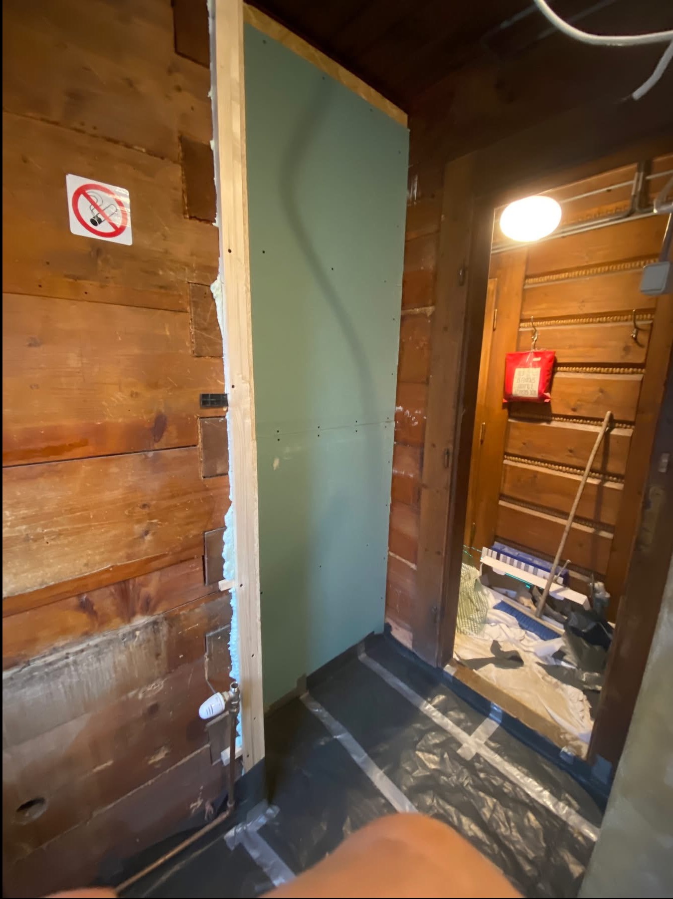 Montaż płyty karton gips na drewnianej ścianie. Widoczne elementy konstrukcyjne i izolacja. Podłoga zabezpieczona folią. W tle drewniane drzwi i pomieszczenie.
